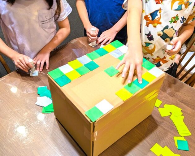 子どもがダンボール箱におりがみを貼っている